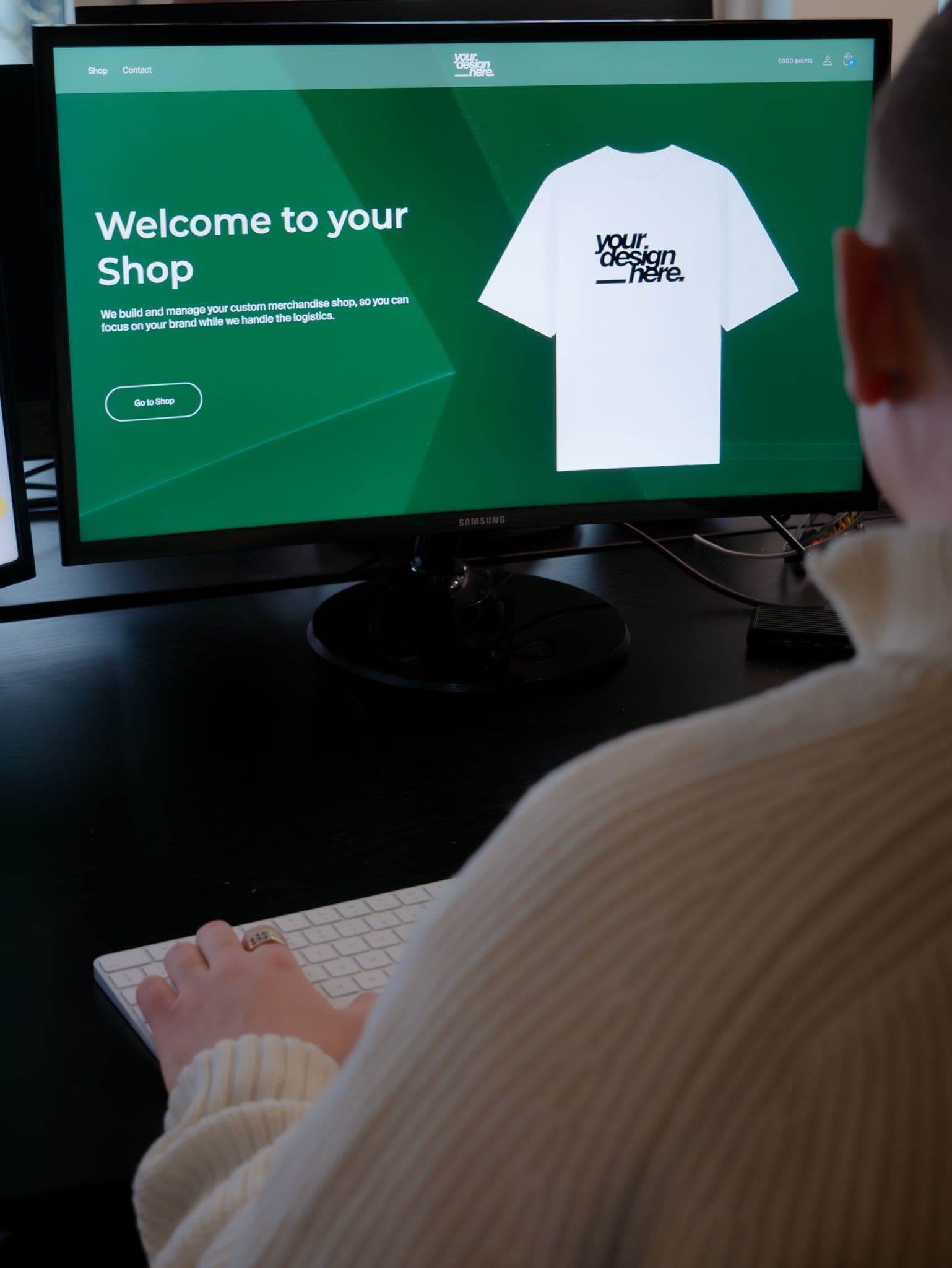 A person at a desk views a computer screen showing a custom merchandise shop website featuring a t-shirt mockup.