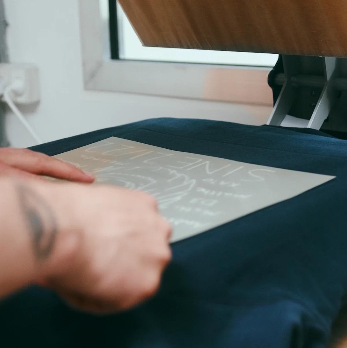 Hands placing a white transfer sheet with a design onto a dark blue t-shirt on a heat press.