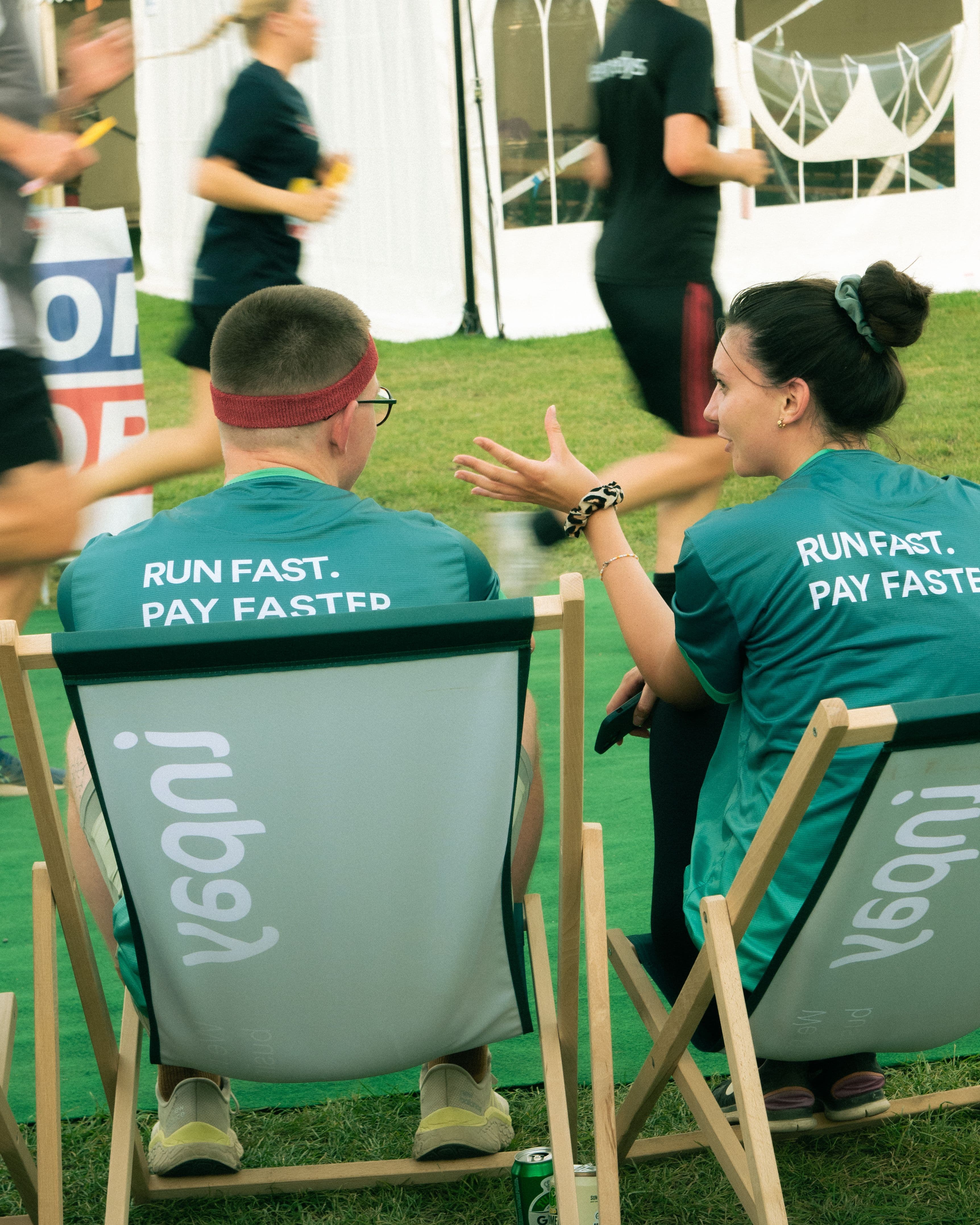 Two people in matching "RUN FAST. PAY FASTED" t-shirts sit in deck chairs at a race, with the woman gesturing as she speaks to the man.