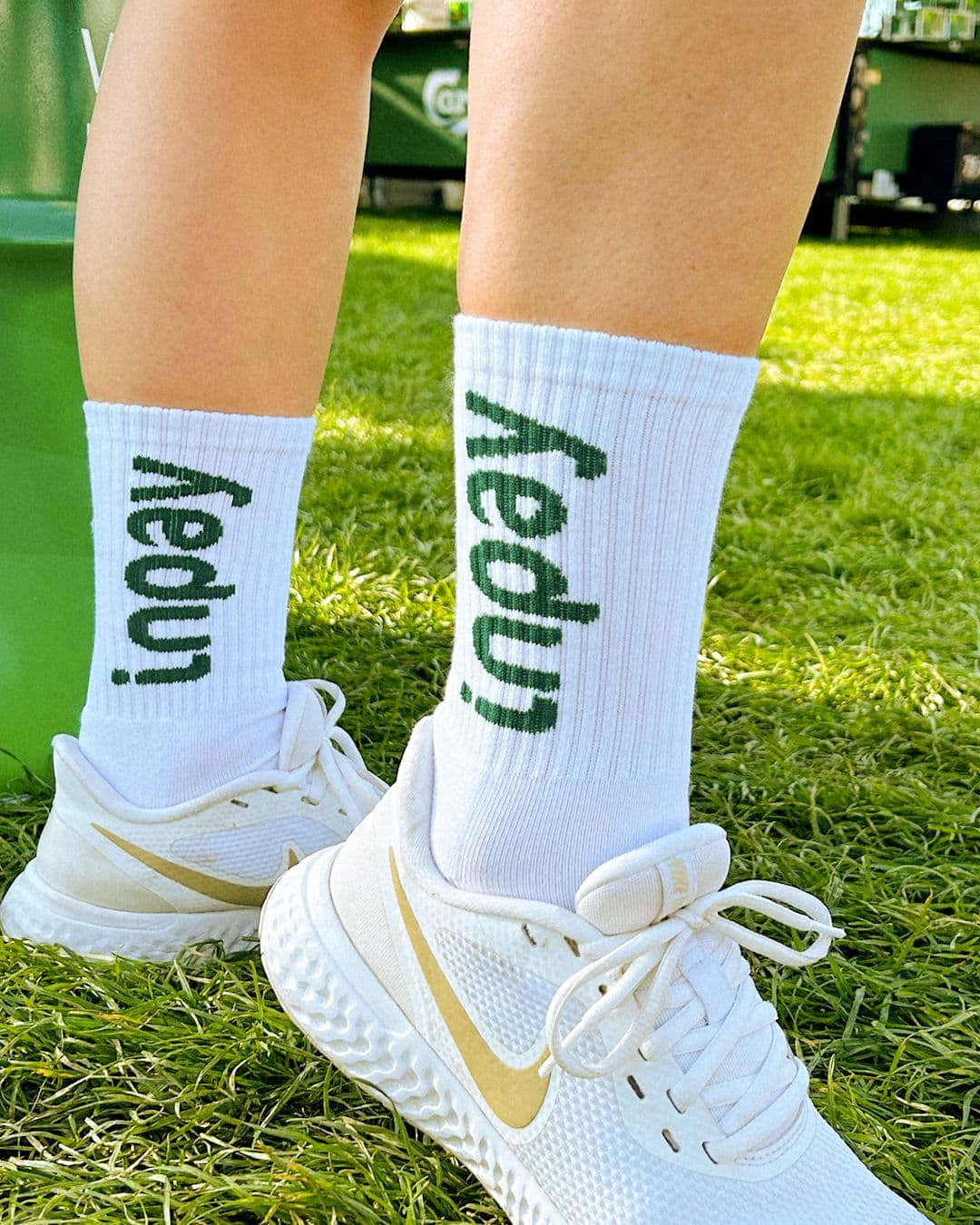 A person's legs wearing white socks with "inpay" in green, and white Nike sneakers with gold swooshes, standing on grass.