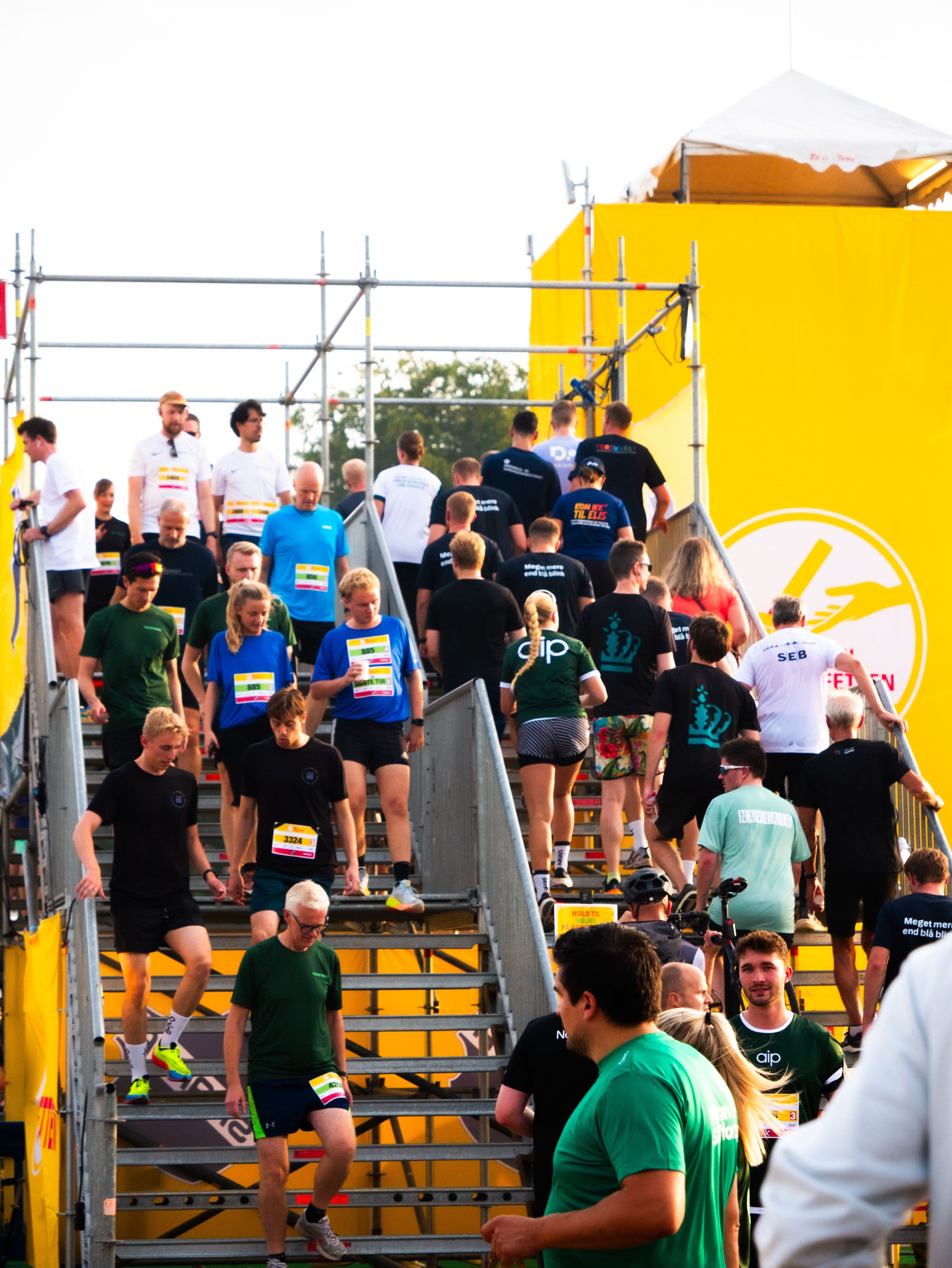 A large group of people in athletic wear and race bibs walk up and down a temporary metal staircase at an outdoor event with a large yellow structure.